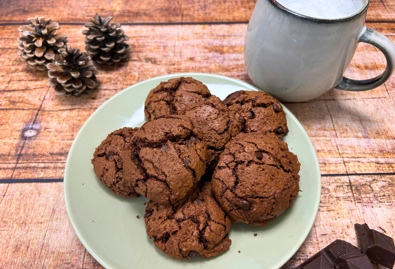 Espresso-Brownie-Cookies auf Porzellanteller mit Emaillebecher, Tannenzapfen und Schokolade auf Holztisch