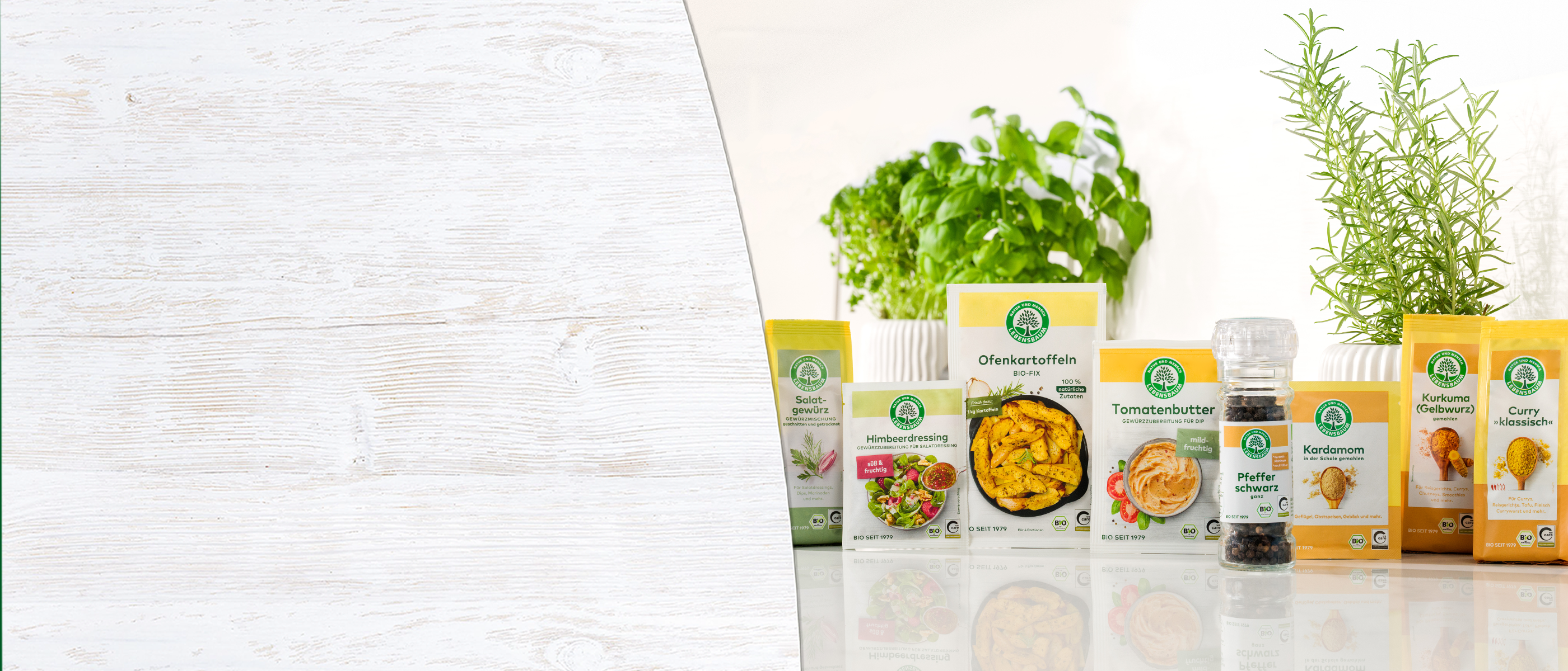 Sortiment von Bio-Gewürzen und Gewürzprodukten mit gelben Packungen, frischen Kräutern und Glasgefäßen auf weißem Holzuntergrund.