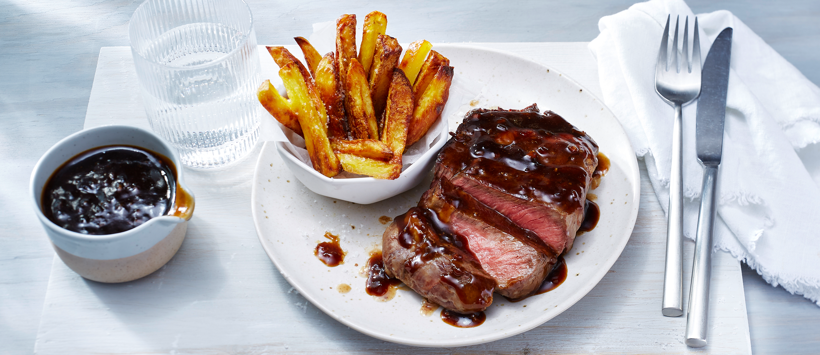 Steak mit Bratensauce und Pommes auf einem Teller, daneben Besteck und ein Glas Wasser