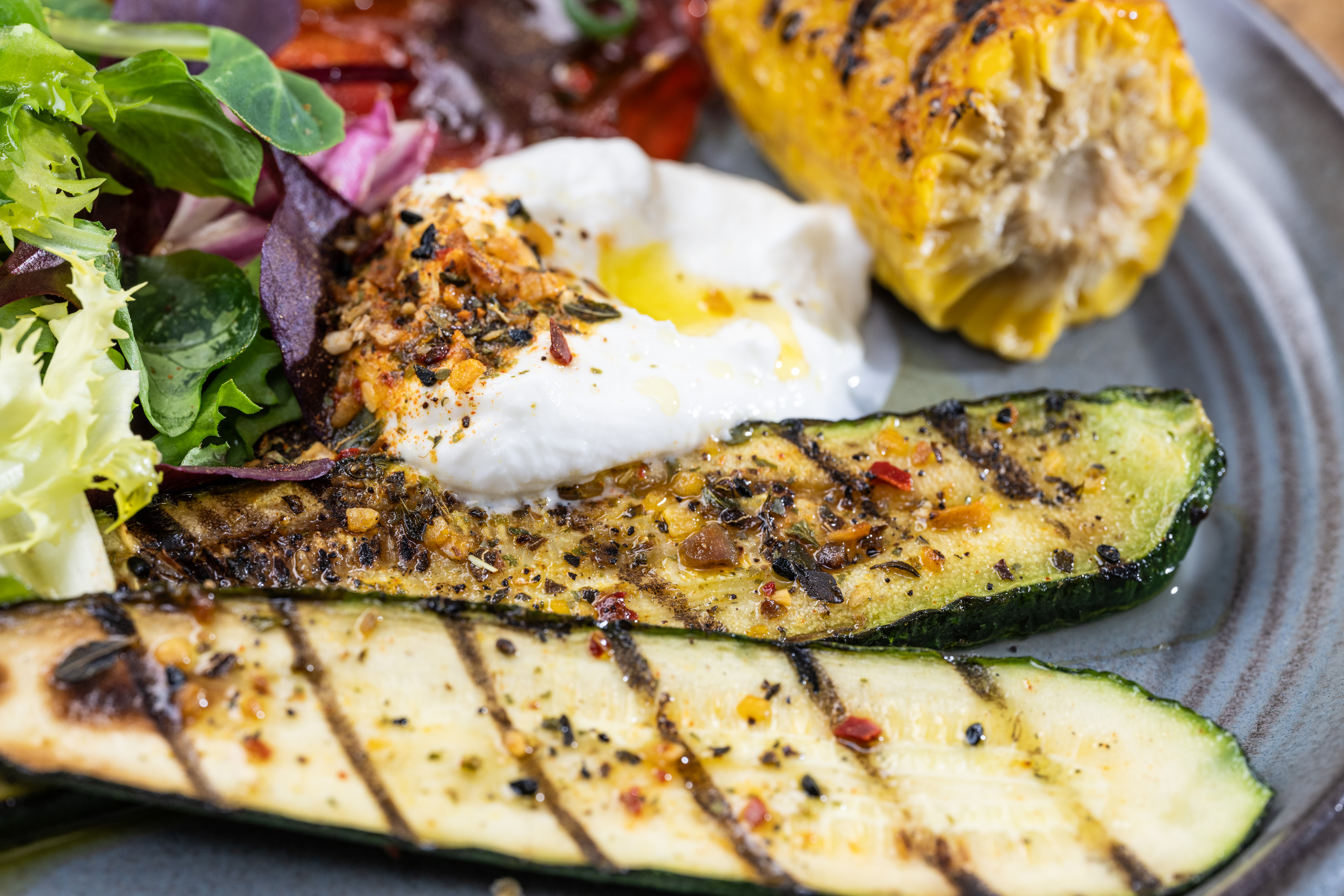 Gegrilltes Gemüse mit Gewürzmischung: Zucchini und Mais mit schwarzen Körnern, Sauerrahm und frischem Salat auf grauem Teller.