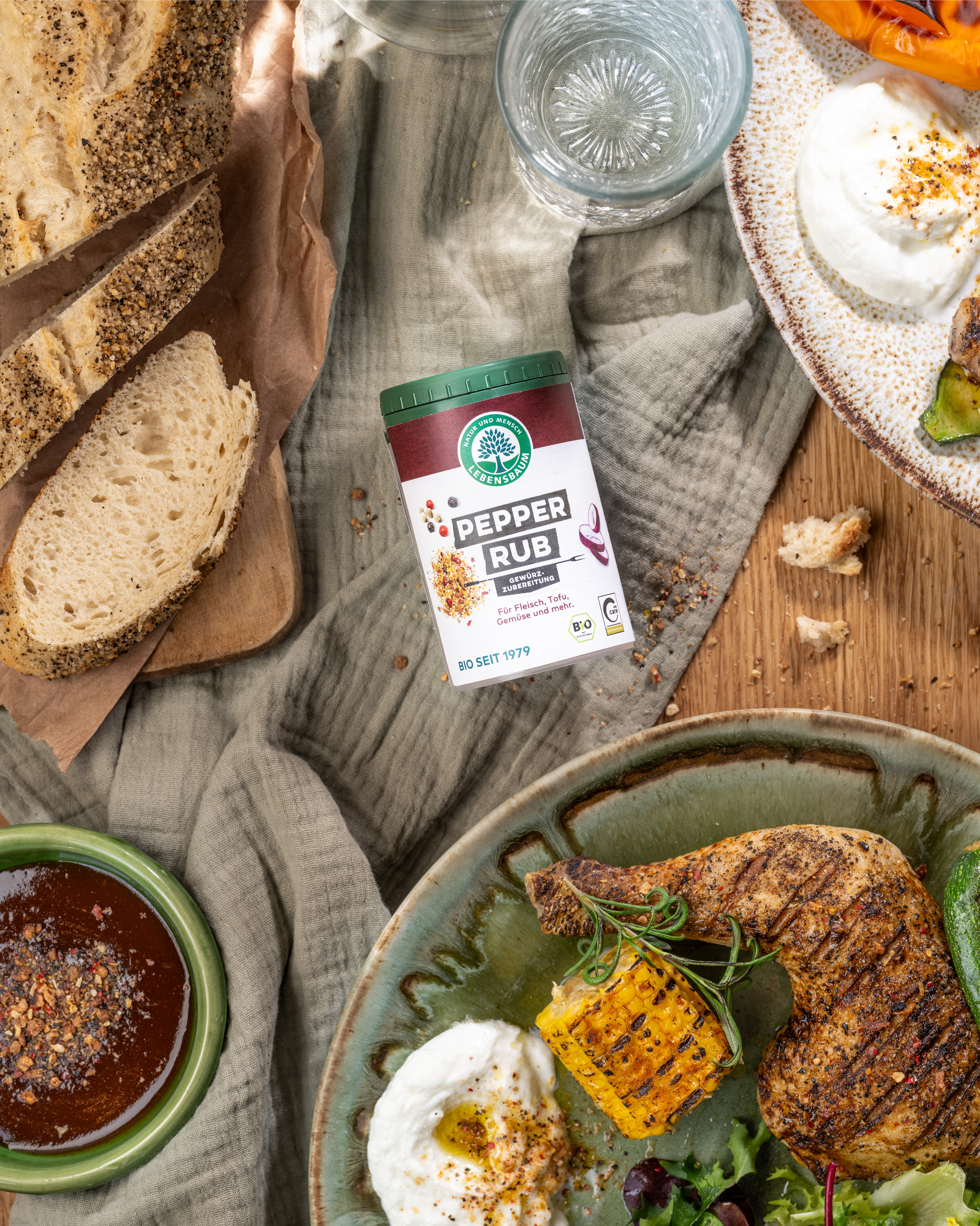 Bio-Gewürzmischung Pepper Rub in rot-weißer Verpackung auf grauem Tuch, umgeben von Brot, gegrilltem Fleisch mit Gewürzkruste, Mais und Sauerrahm.