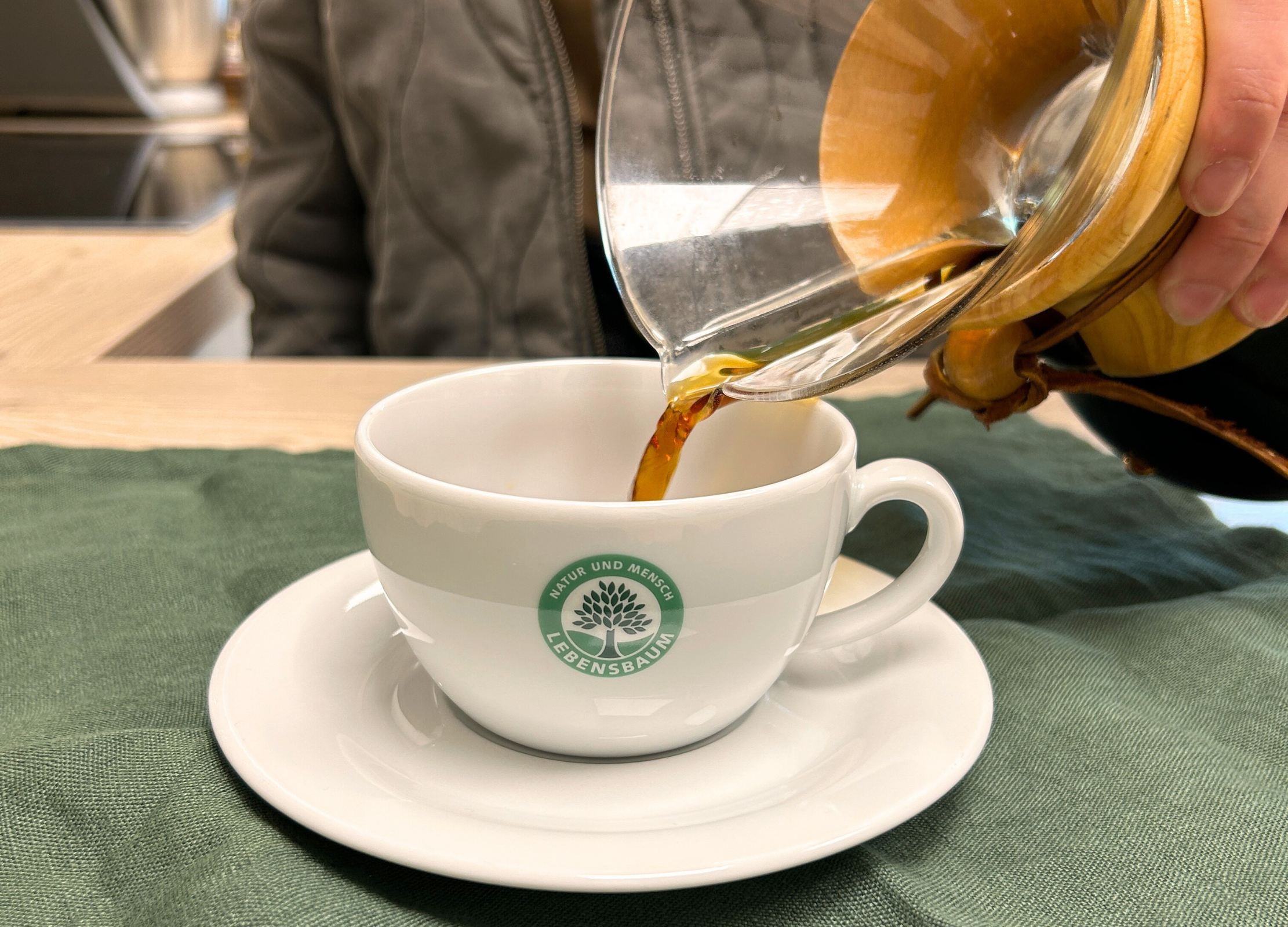 Frisch gebrühter Kaffee wird aus einer Chemex-Karaffe in eine Tasse mit Lebensbaum-Logo gegossen