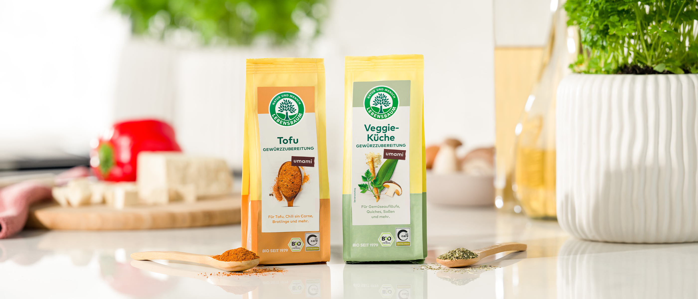 Zwei Bio-Gewürzmischungen der Marke Lebensbaum: Veggie-Tofu in gelber Packung mit orangefarbenem Etikett und Veggie-Küche in grüner Packung, daneben Holzlöffel mit Gewürzen auf heller Küchenarbeitsplatte, grüne Pflanzen im Hintergrund.