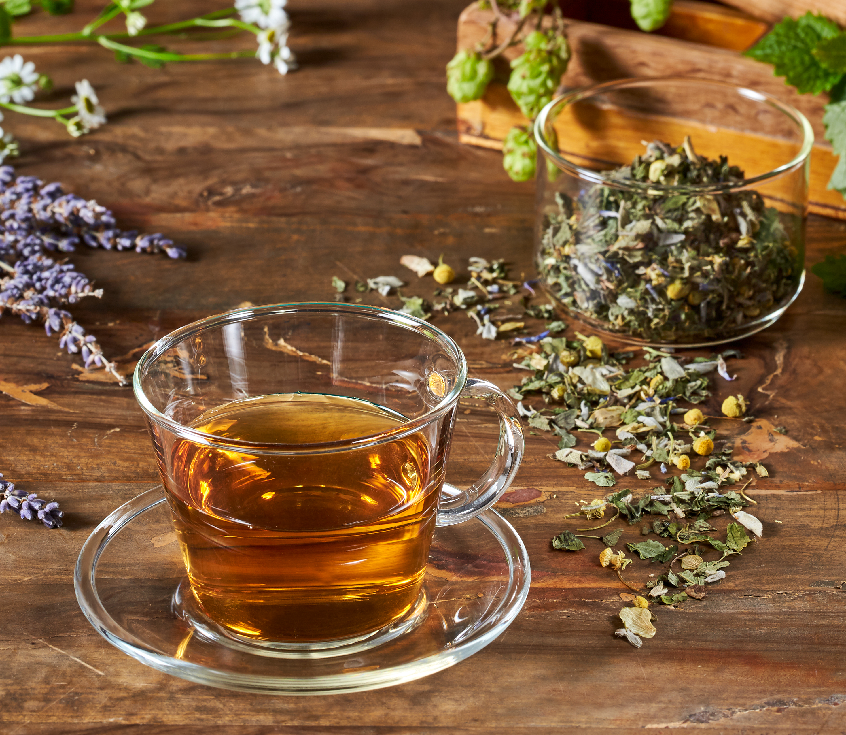 Tasse goldgelber Kräutertee mit Glastasse und Untertasse auf Holztisch, umgeben von getrockneten Kräutern, Kamille, Lavendel und Hopfen.