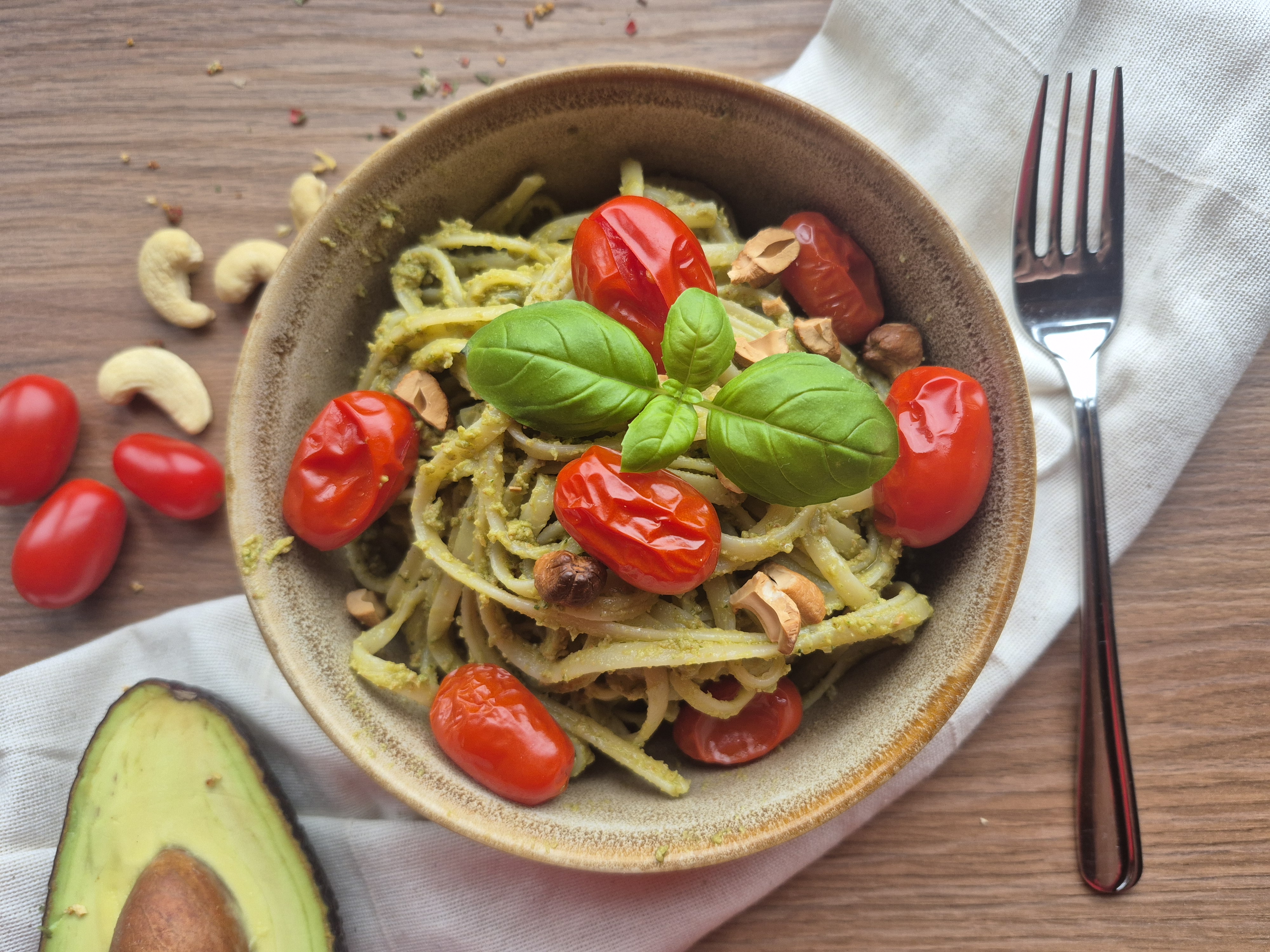 Grüne Pasta mit Basilikum-Pesto in Tonschale, garniert mit roten Tomaten, Cashewkernen und frischem Basilikum, serviert mit Avocado.