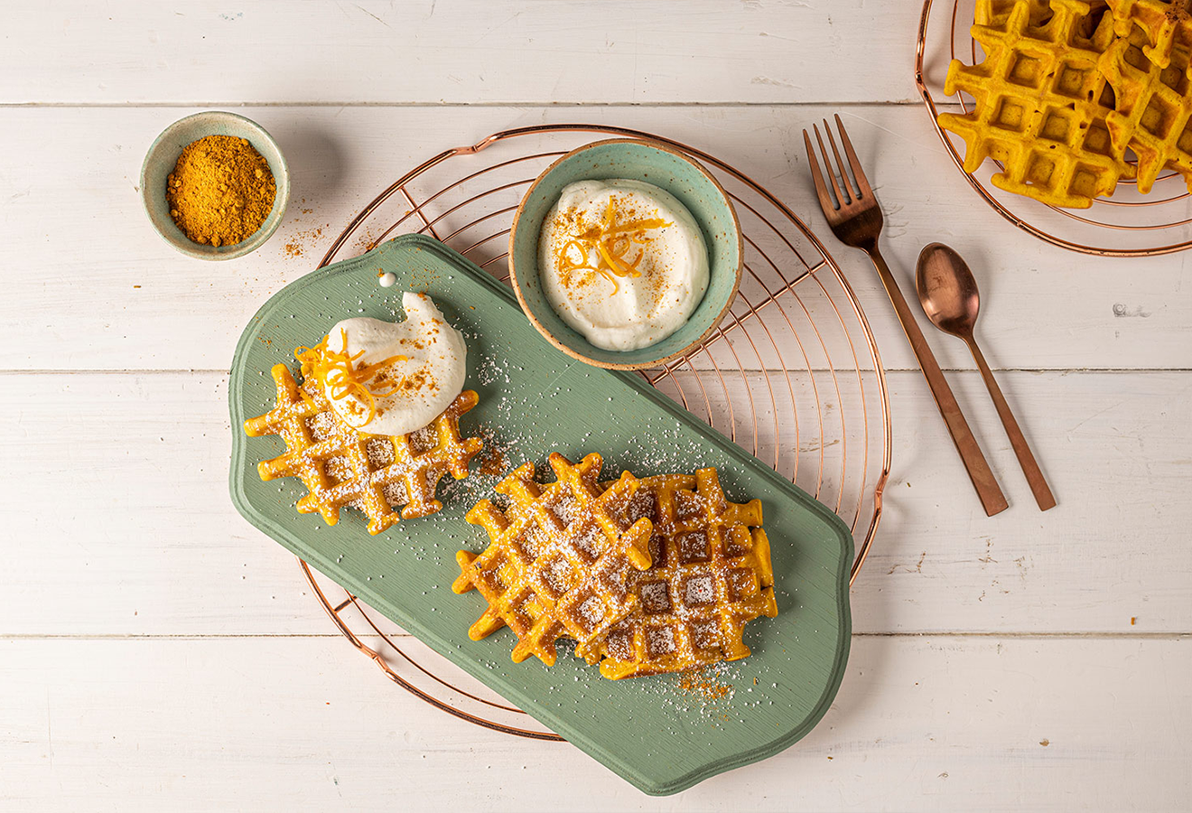 Kürbis-Waffeln mit Puderzucker und Kurkuma auf grünem Teller, serviert mit Sahne-Dip und orangefarbenen Waffeln im Hintergrund.
