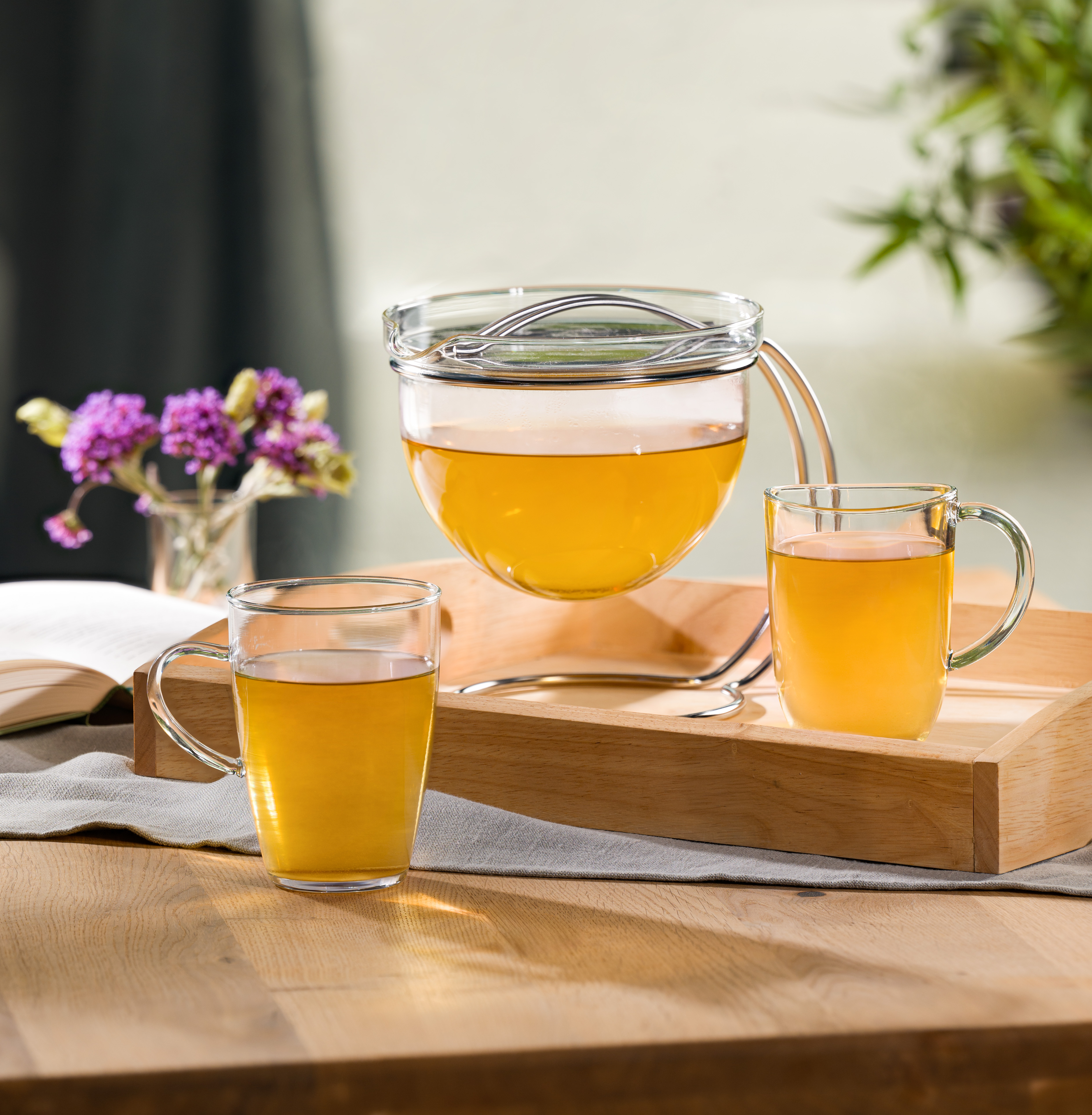Teezubereitung mit Glaskanne und zwei Tassen goldenem Tee auf Holztablett, pink blühende Blume und aufgeschlagenes Buch im Hintergrund