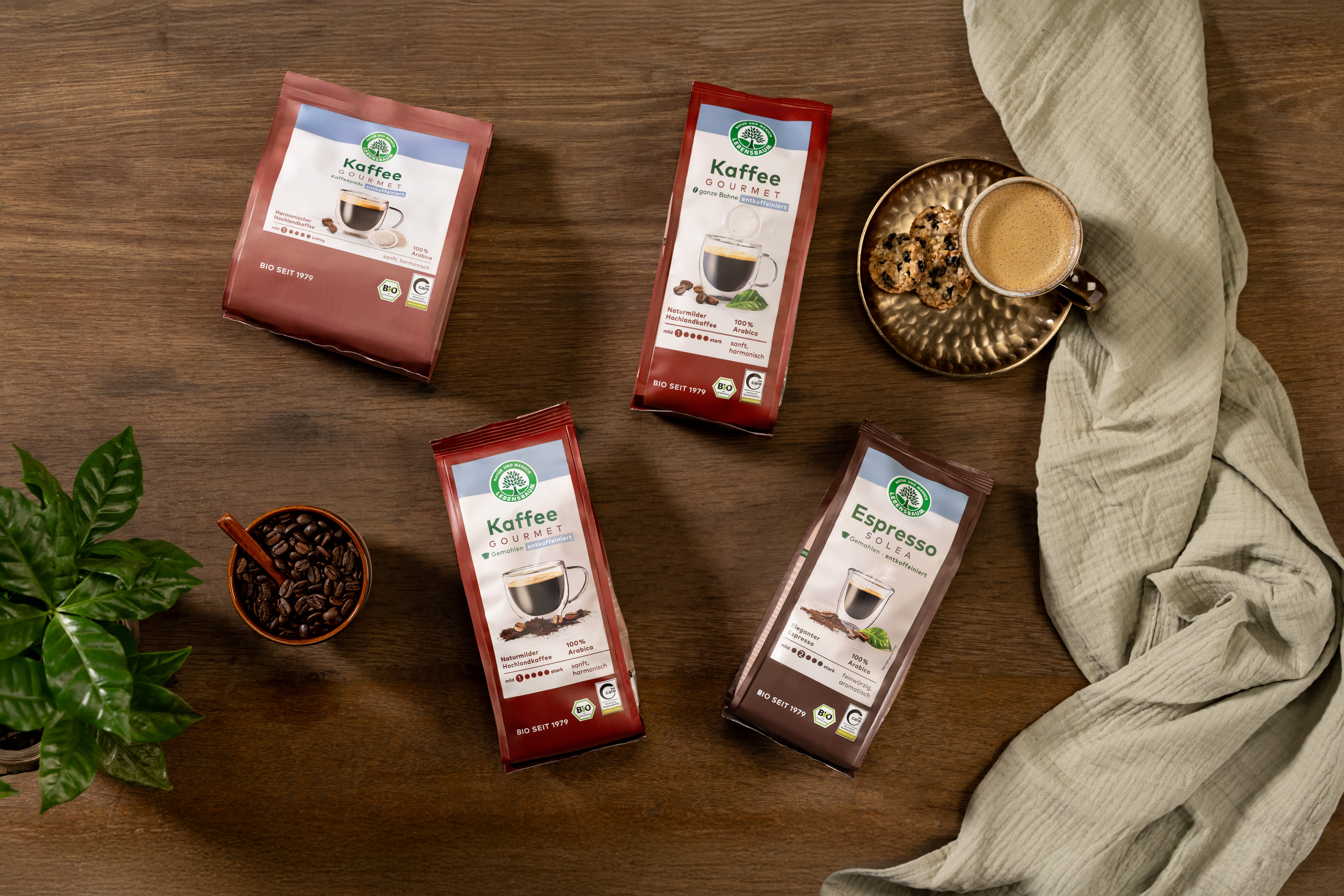 Lebensbaum Bio-Kaffee Sortiment mit roten und braunen Verpackungen auf Holztisch, daneben Kaffeetasse mit Schaum, Kekse, Kaffeebohnen in Schale und grüne Pflanze.