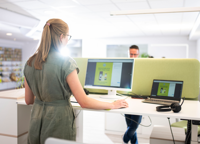Frau mit Brille am Schreibtisch mit Monitor und Laptop in modernem Büro, grüne Trennwand, Kollege im Hintergrund.