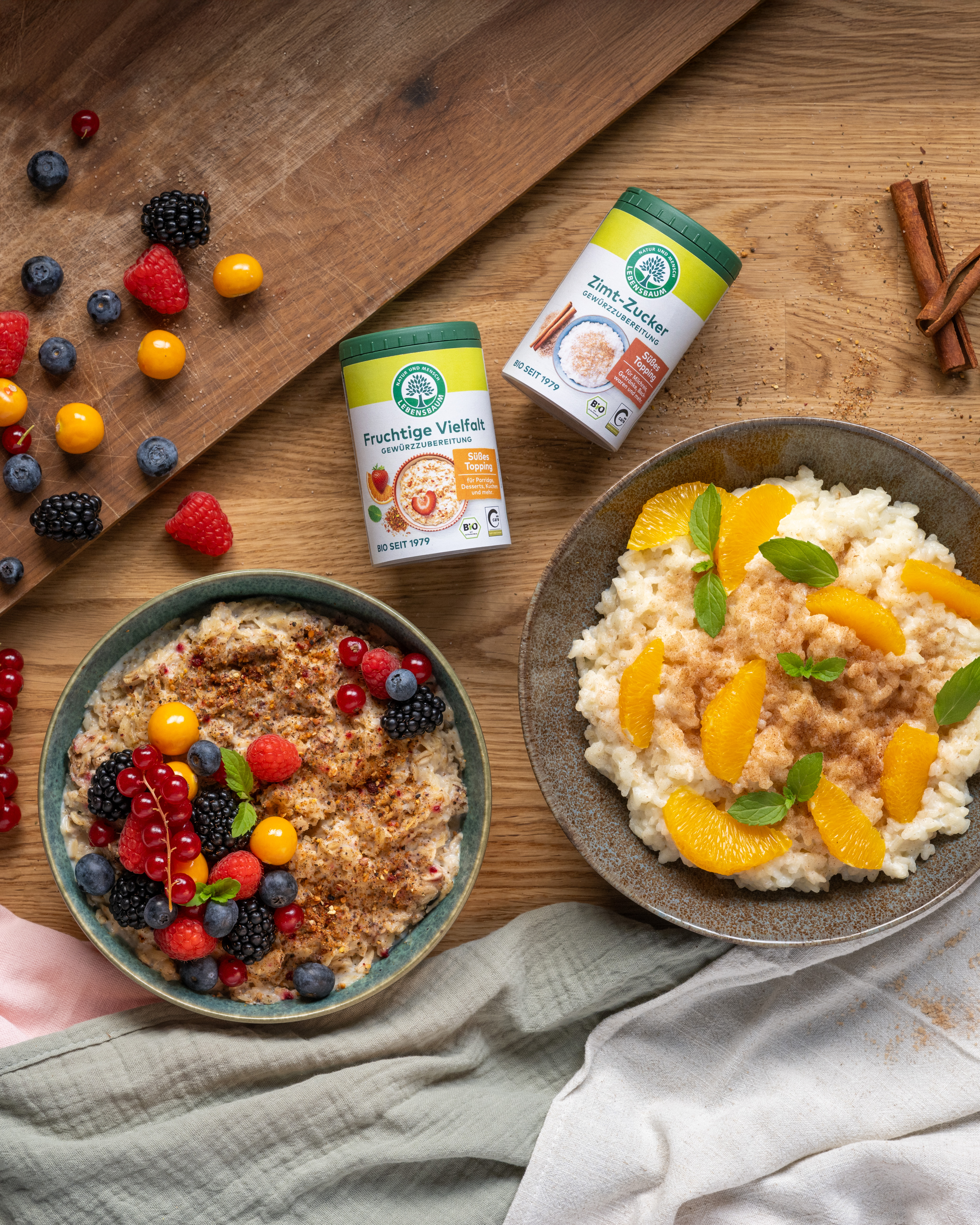 Zwei Frühstücksbowls mit Müsli, Beeren und Orangenscheiben neben Gewürzprodukten "Fruchtige Vielfalt" und "Zimt-Zucker" auf Holztisch mit Zimtstangen
