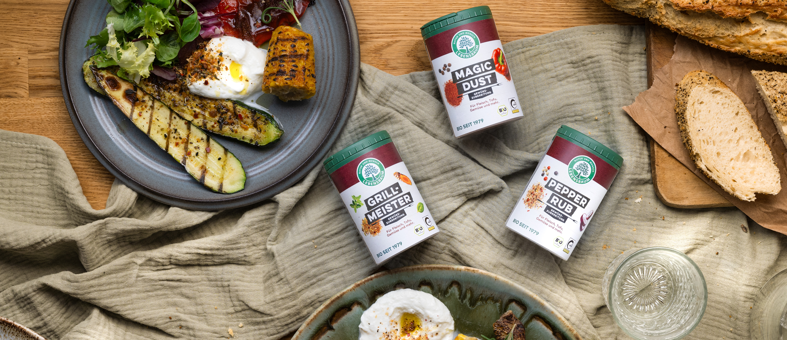 Gourmet-Gewürze Magic Dust, Grillmeister und Pepper Rub neben gegrilltem Gemüse, Brot und Eierspeisen auf Holztisch
