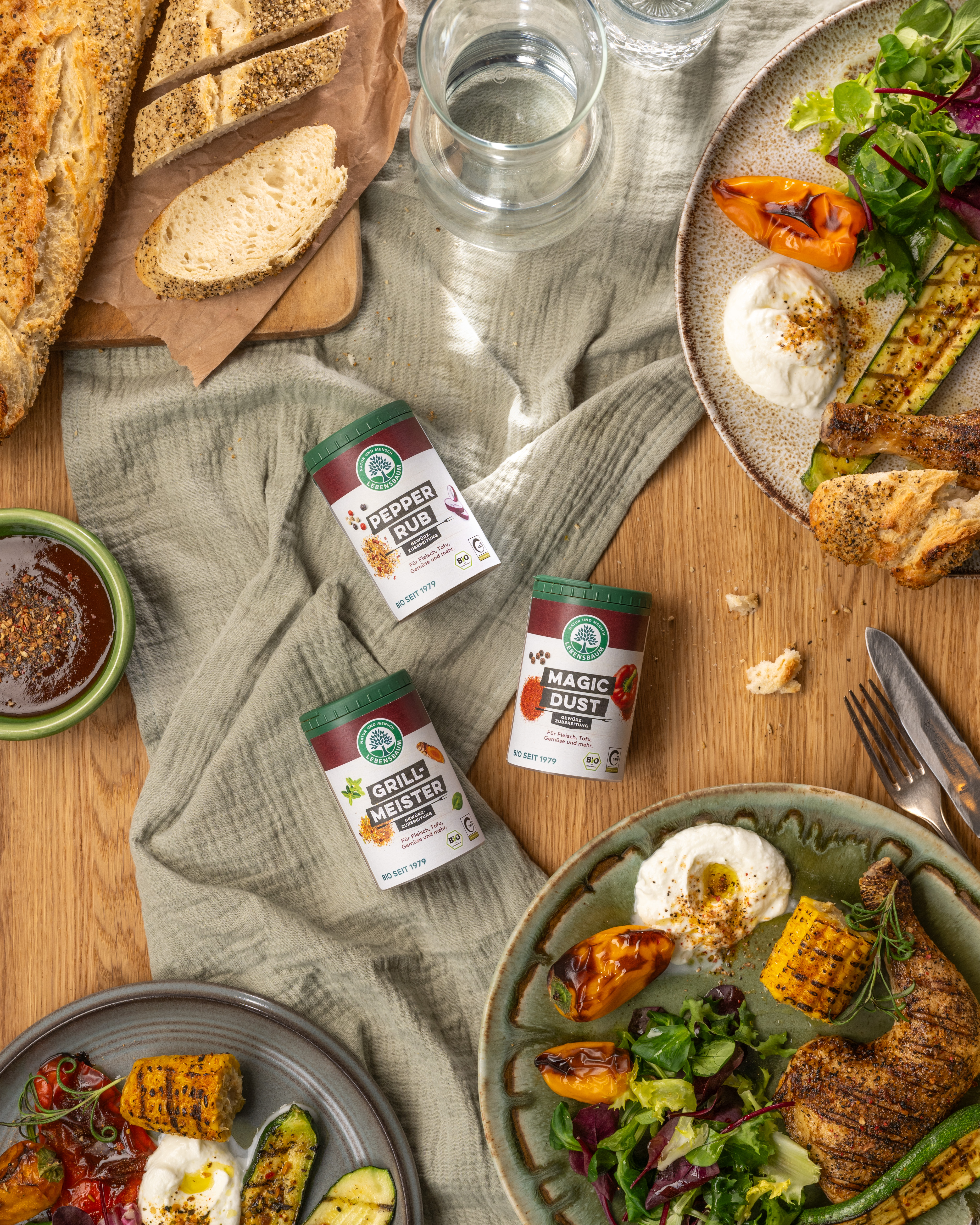 Pepper Rub, Grill Meister und Magic Dust Gewürzmischungen auf Holztisch mit Grillgemüse, Toast und Teller mit gegrilltem Fleisch