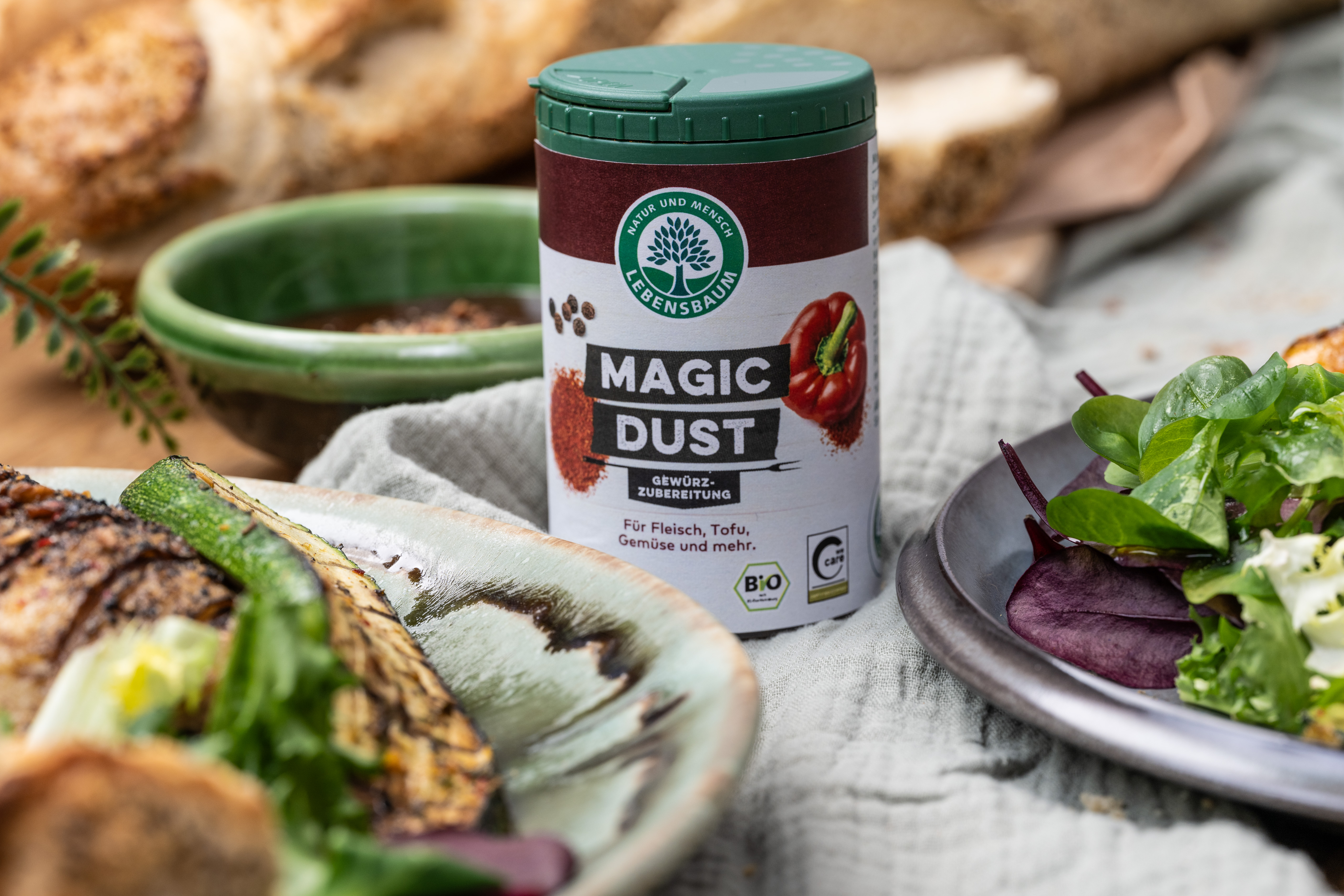 Magic Dust Gewürzzubereitung in Streudose umgeben von frischem Gemüse, Brot und Kräutern auf Holzuntergrund.