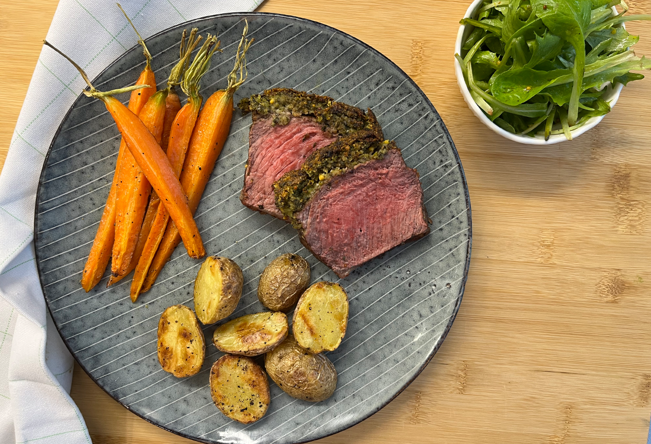 Angerichtetes Gericht auf grauem Teller: medium-rare Fleisch mit Umami-Gewürzkruste, roastete Kartoffeln, orange Möhren und frischem Rucola.