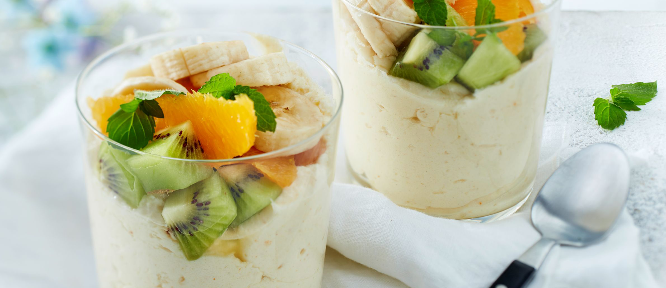 Zwei Dessertgläser mit Creme, garniert mit Bananen, Kiwi, Orange und Minze