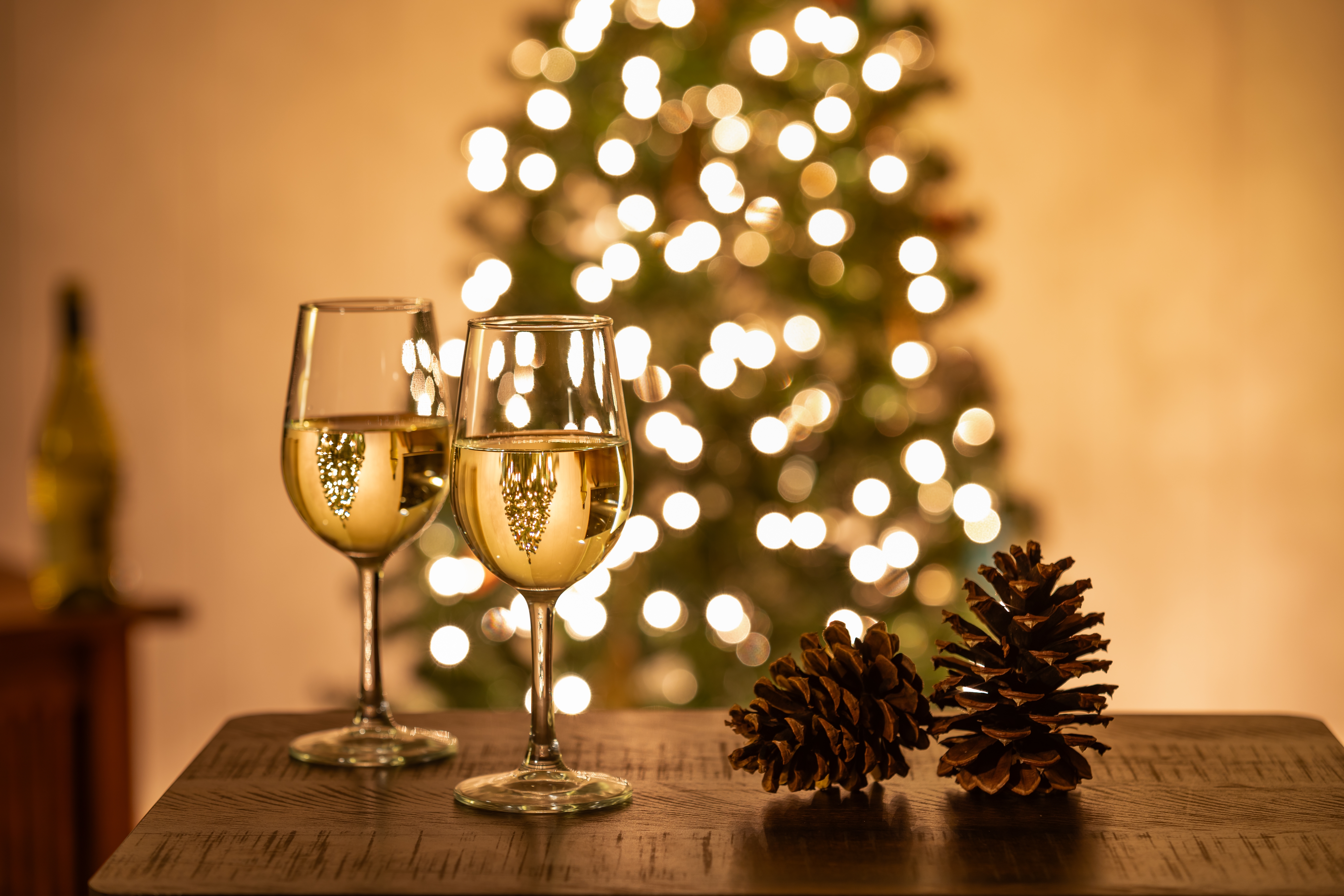 Zwei Gläser Weißwein mit Blasen vor festlich illuminiertem Weihnachtsbaum und Tannenzapfen