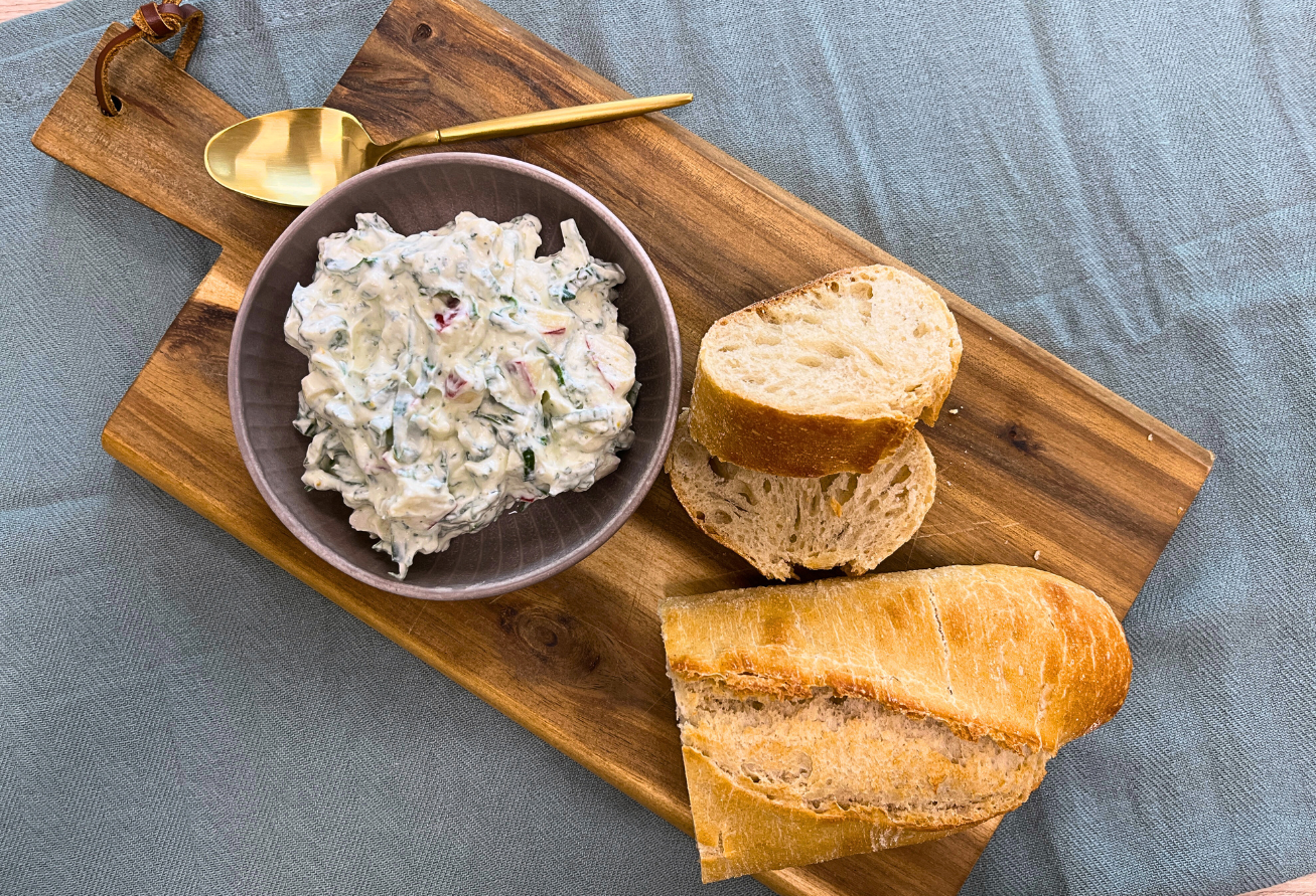 Brotaufstrich mit Lauchgrün in grauer Schale auf Holzbrett, serviert mit Brotscheiben und goldenem Löffel