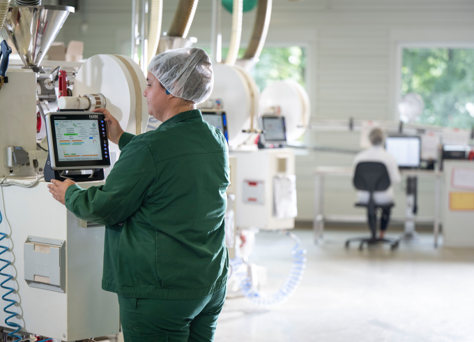 Mitarbeiterin in grüner Arbeitskleidung kontrolliert Laborgerät auf Touchscreen in moderner Produktionsstätte.