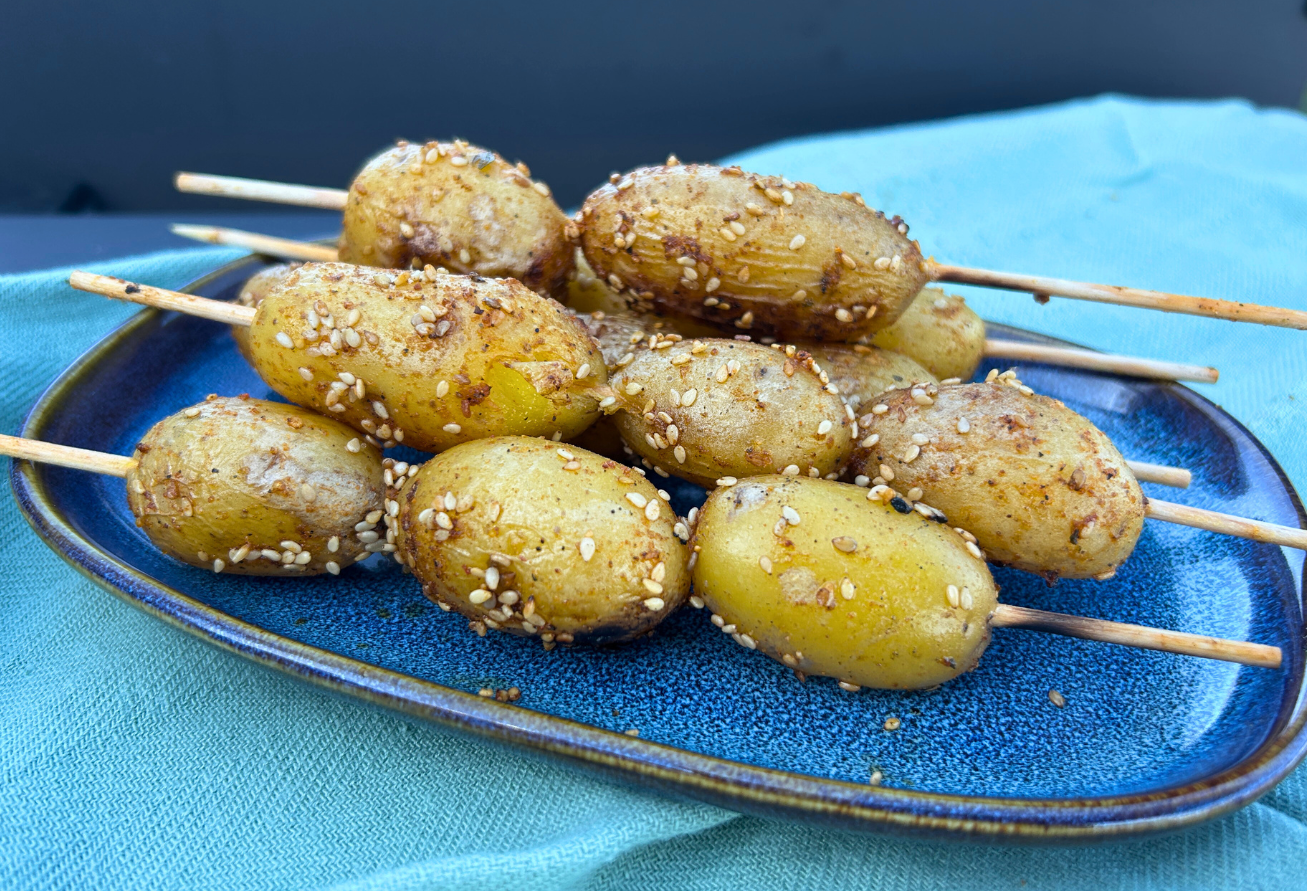 Gegrillte Kartoffelspieße mit Sesam und Gewürzen auf blauem Teller, serviert auf hellblauem Untergrund.