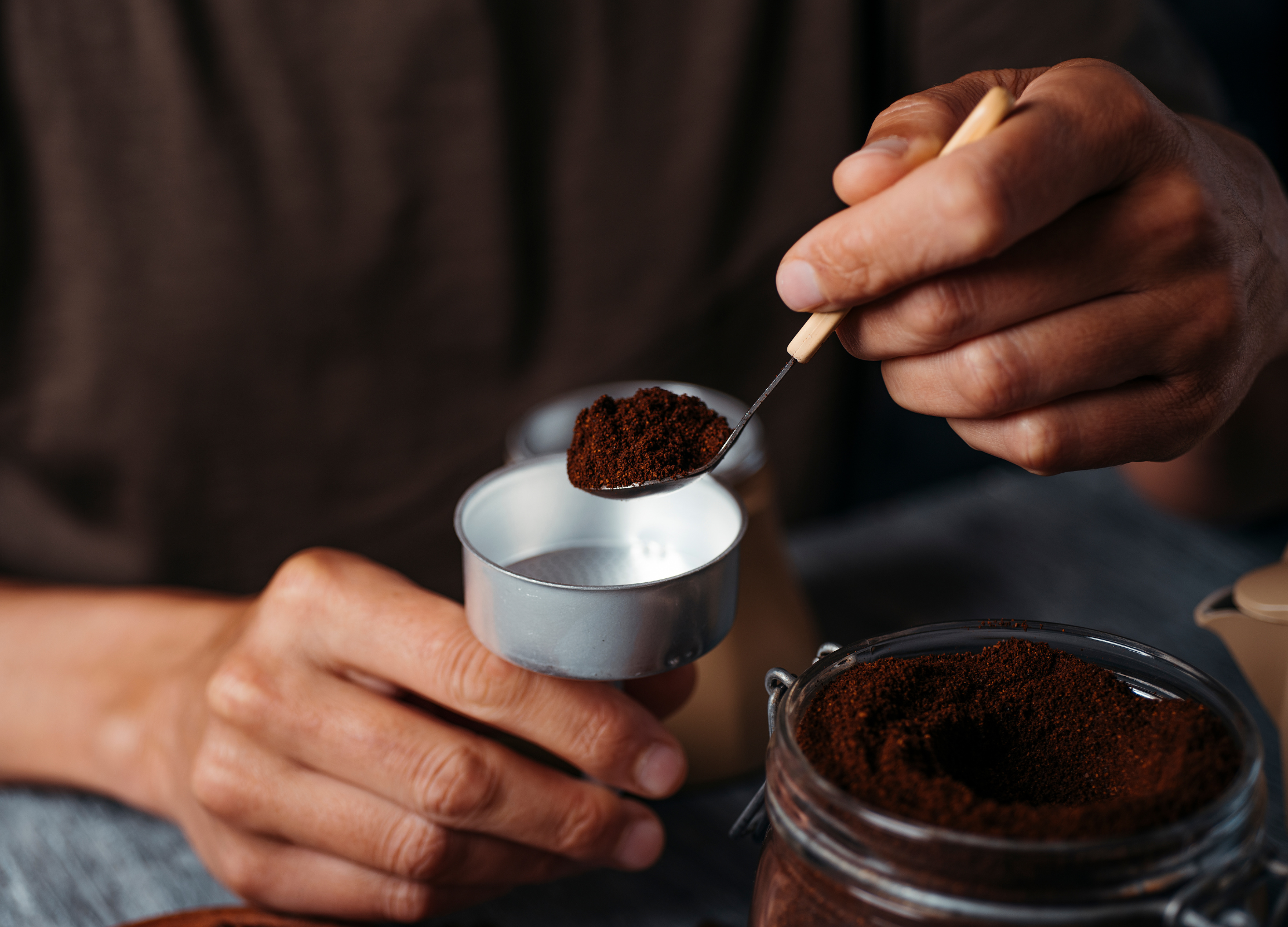 Person füllt gemahlenen Kaffee mit einem Löffel aus einem Glasbehälter in einen silbernen Siebträgereinsatz