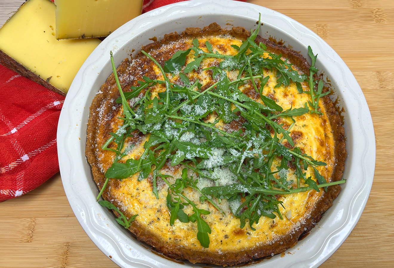 Käse-Quiche in weißer Auflaufform mit goldbrauner Oberfläche, garniert mit frischen Rucola-Blättern und Parmesan, neben gelbem Käse auf Holztisch.