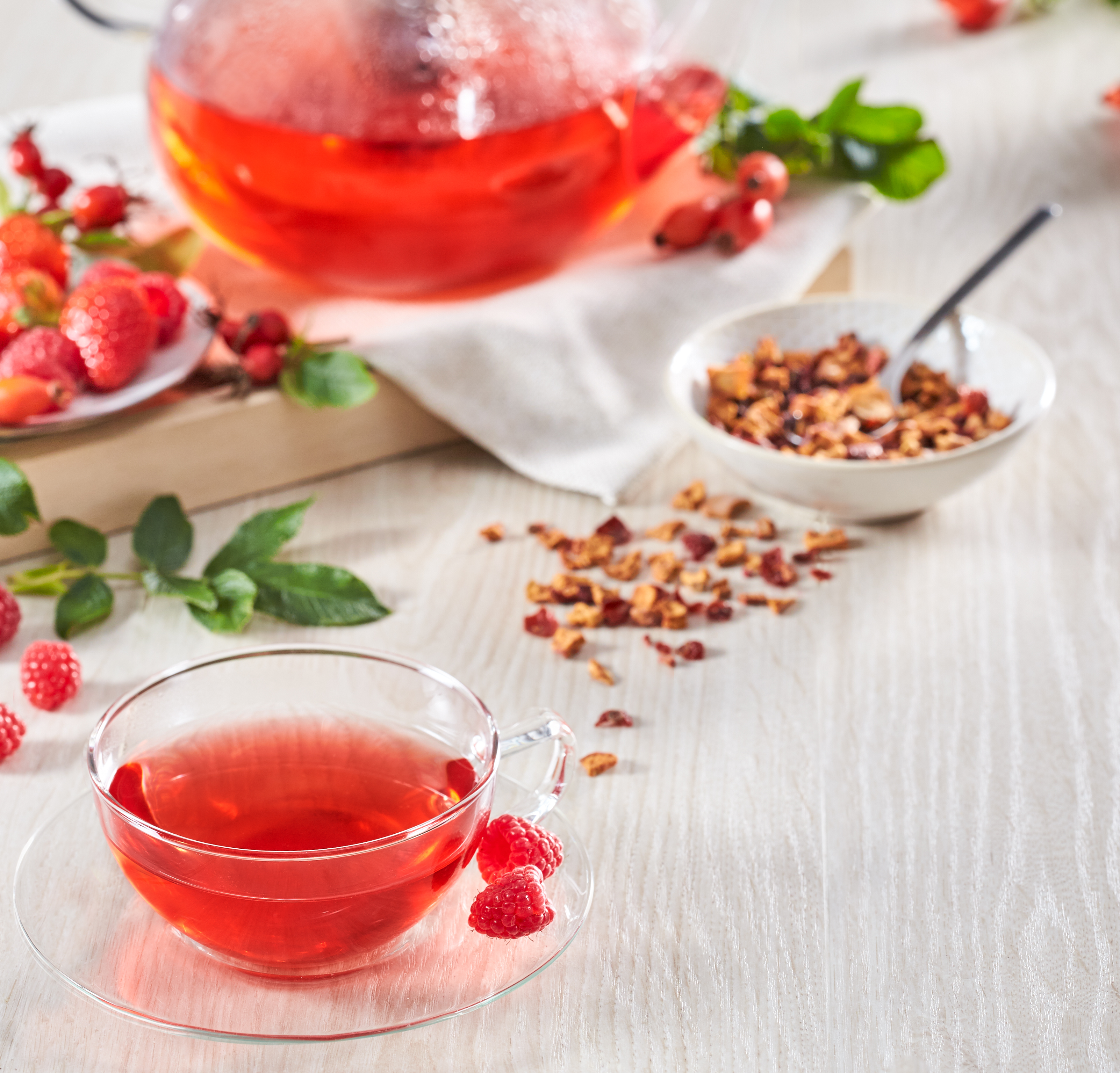 Rote Beerentee-Zubereitung mit frischen Himbeeren, getrockneten Beeren und Früchtekräutern auf hellem Holztisch.