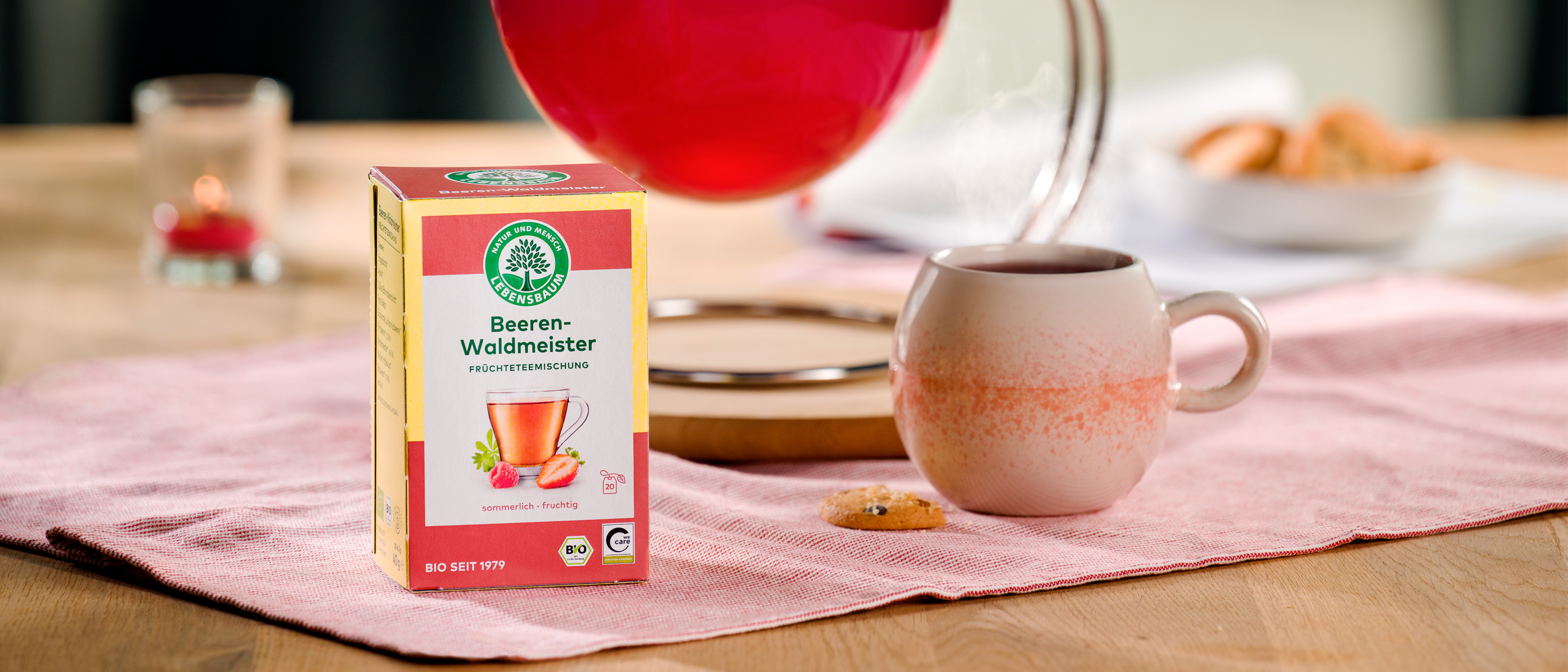 Packung Lebensbaum Beeren-Waldmeister-Tee steht neben einer Tasse, in die frisch aufgebrühter Tee aus einer Glaskanne gegossen wird