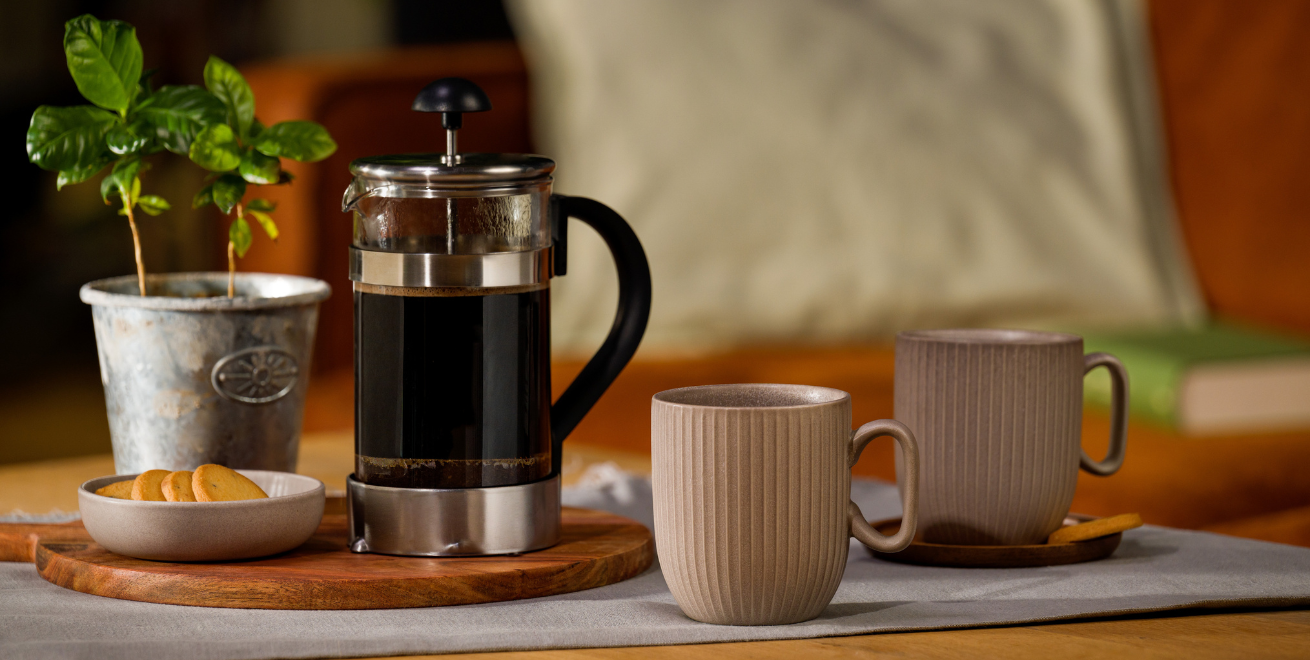 Eine mit Kaffee gefüllte French Press steht neben einem Teller mit Keksen und einer kleinen Pflanze. Daneben stehen zwei Kaffeebecher.