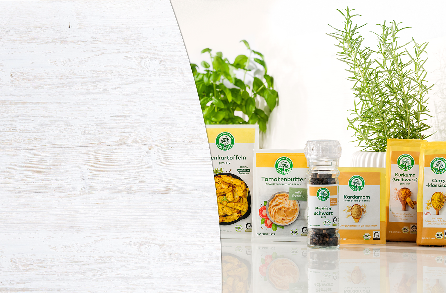 Bio-Gewürze und Kräuter in gelben Verpackungen: Kartoffeln, Tomatenbutter, Pfeffer, Kardamom und Kurkuma, arrangiert mit frischen Kräutern vor weißem Holzhintergrund.