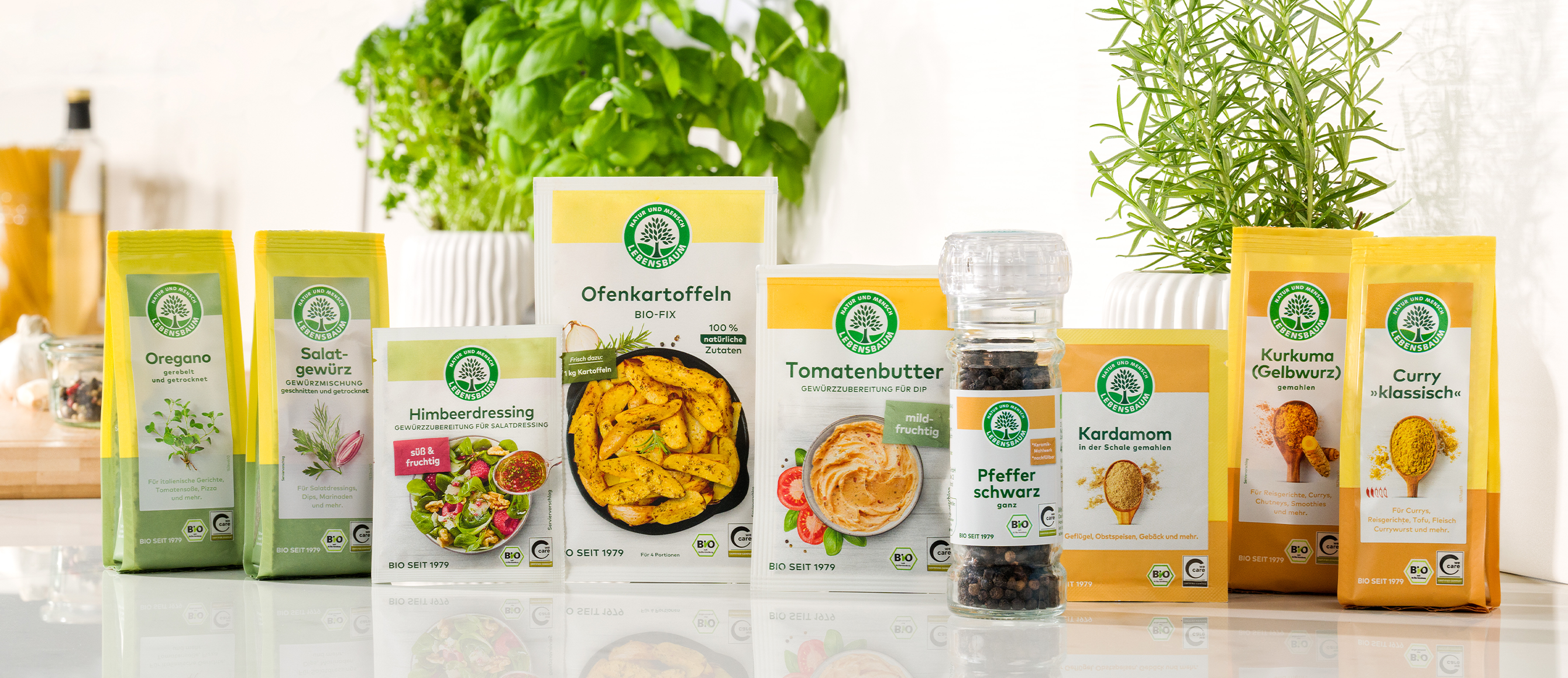Bioprodukt-Sortiment mit Oregano, Salbei-Gewürz, Himbeeressing, Bio-Ofenkartoffeln, Tomatenbutter, schwarzem Pfeffer, Kardamom und Curry-Gewürz in gelb-grünen Verpackungen.