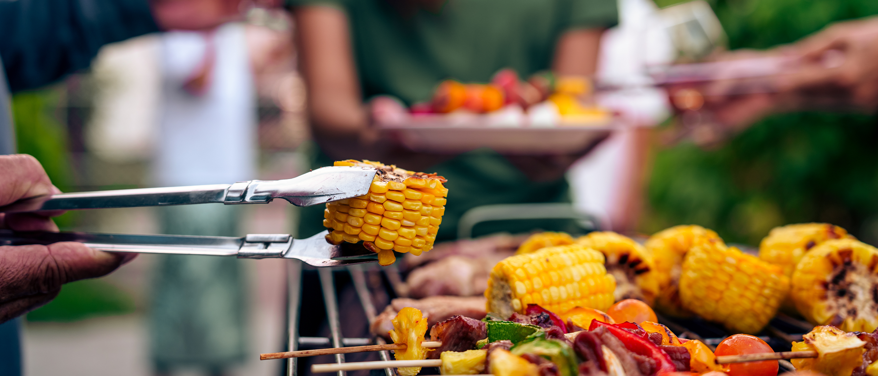 Gegrillter Mais mit Röstmarks auf Zange, im Hintergrund Grillgut und bunte Gemüsespieße bei Gartengrillparty