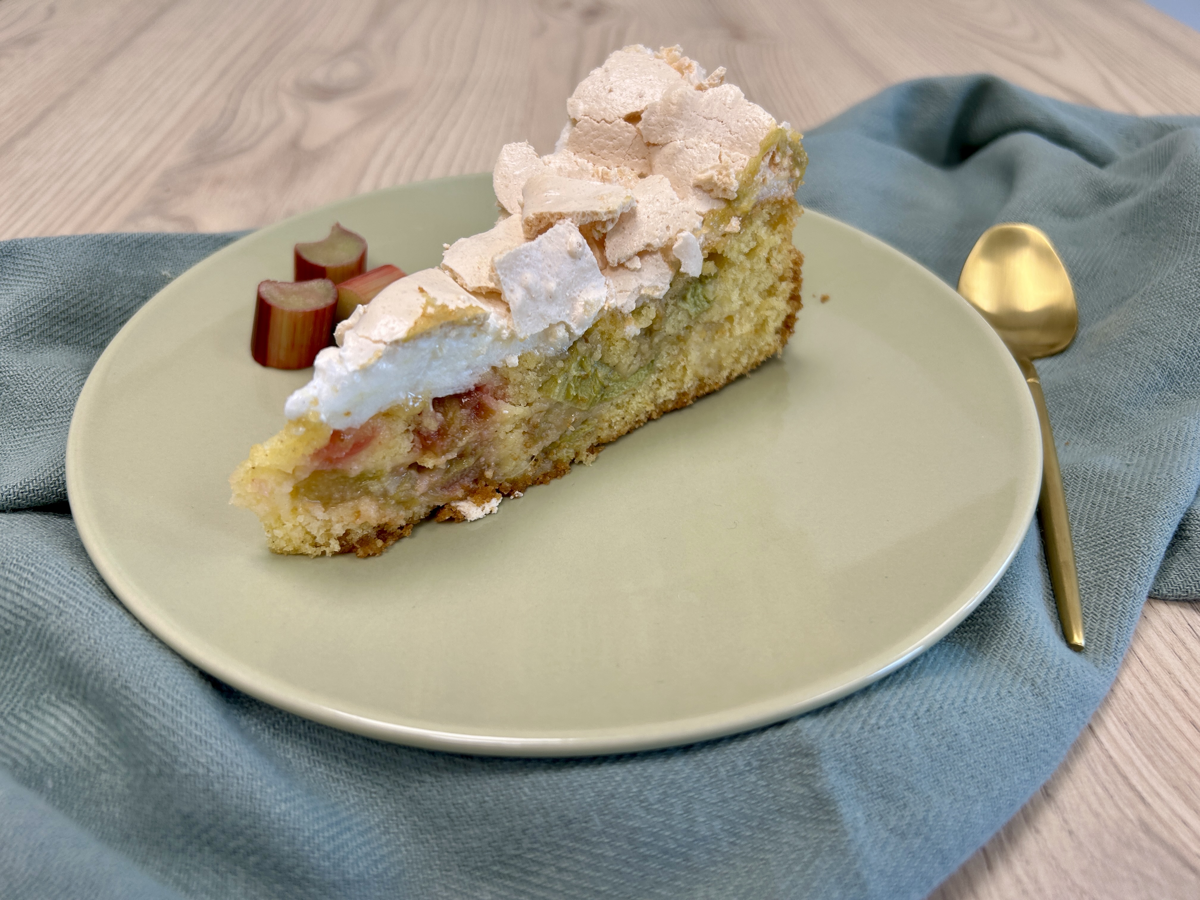 Rhabarberkuchen-Stück auf cremefarbenem Teller mit Baiserhaube, Rhabarberstücken als Garnitur und Goldlöffel.