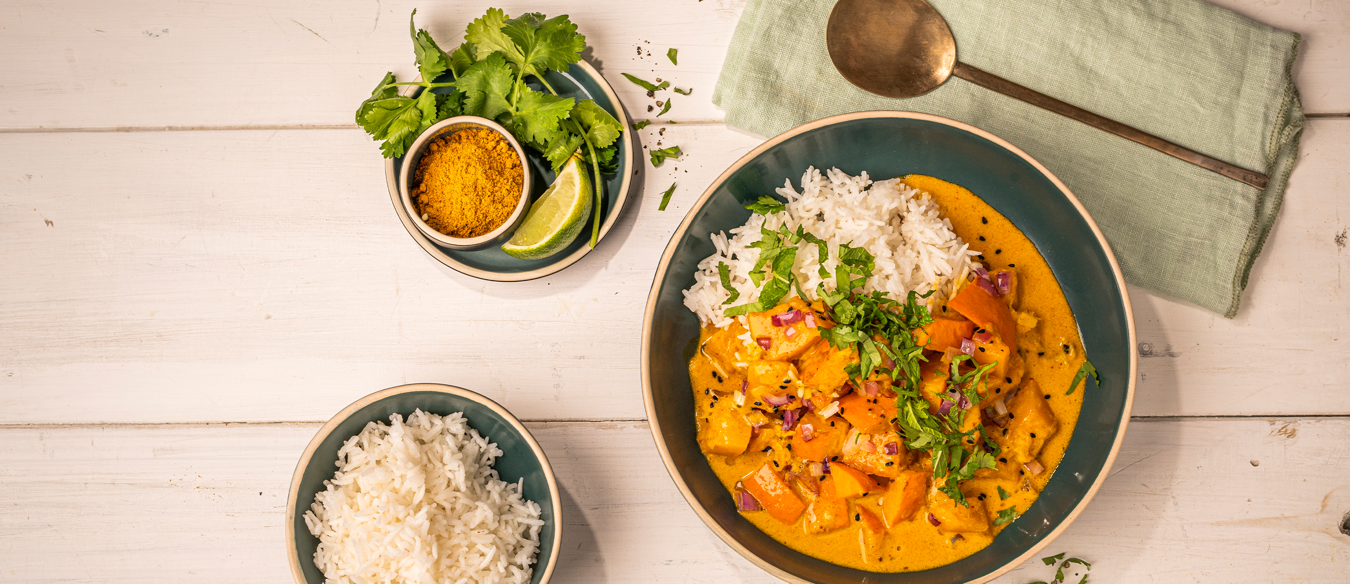 Kürbis-Curry mit Reis, Koriander und Gewürzmischung in grünen Schüsseln auf weißem Holztisch