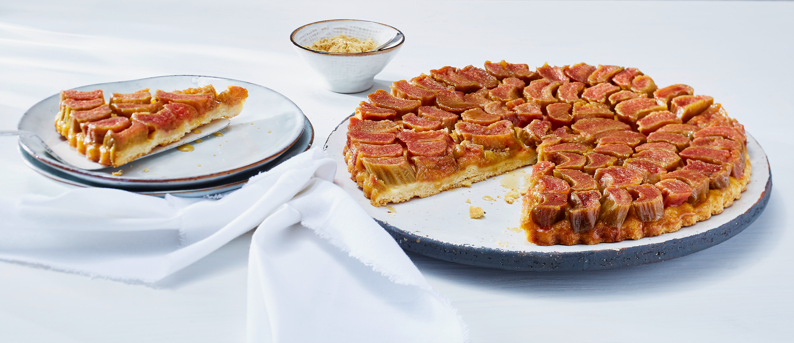 Rhababerkuchen auf einem Kuchentablett, daneben ein weißer Teller mit einem Stück vom Kuchen darauf