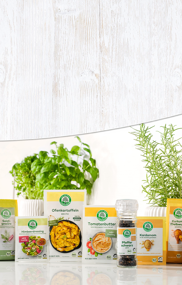 Arrangierte Bio-Gewürzprodukte mit gelben Verpackungen, frischen Kräutern in weißen Töpfen vor Holzwand-Hintergrund.