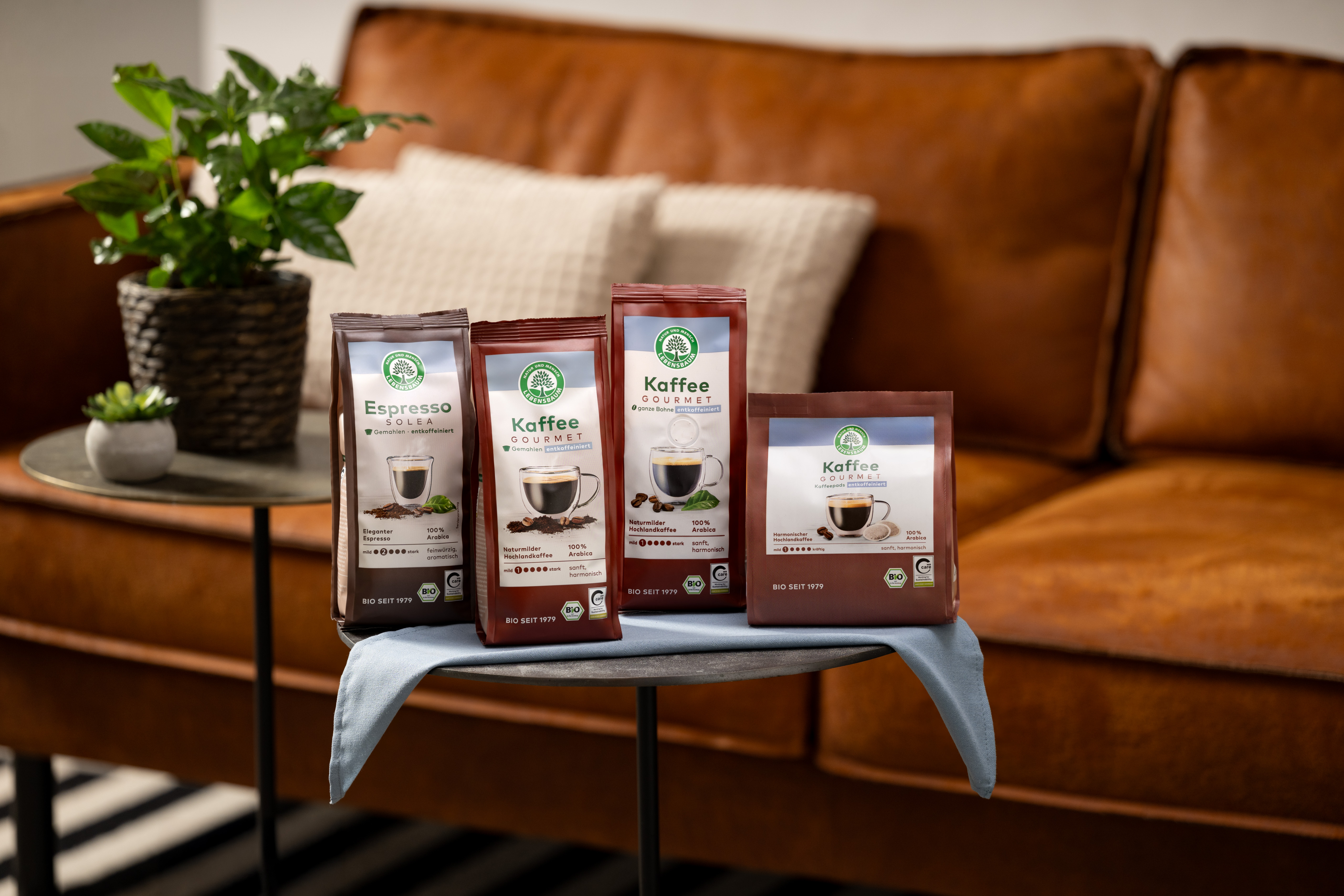 Vier Bio-Kaffeeverpackungen der Sorte Espresso und Gourmet Kaffee mit grünem Siegel auf Couchtisch vor braunem Ledersofa.