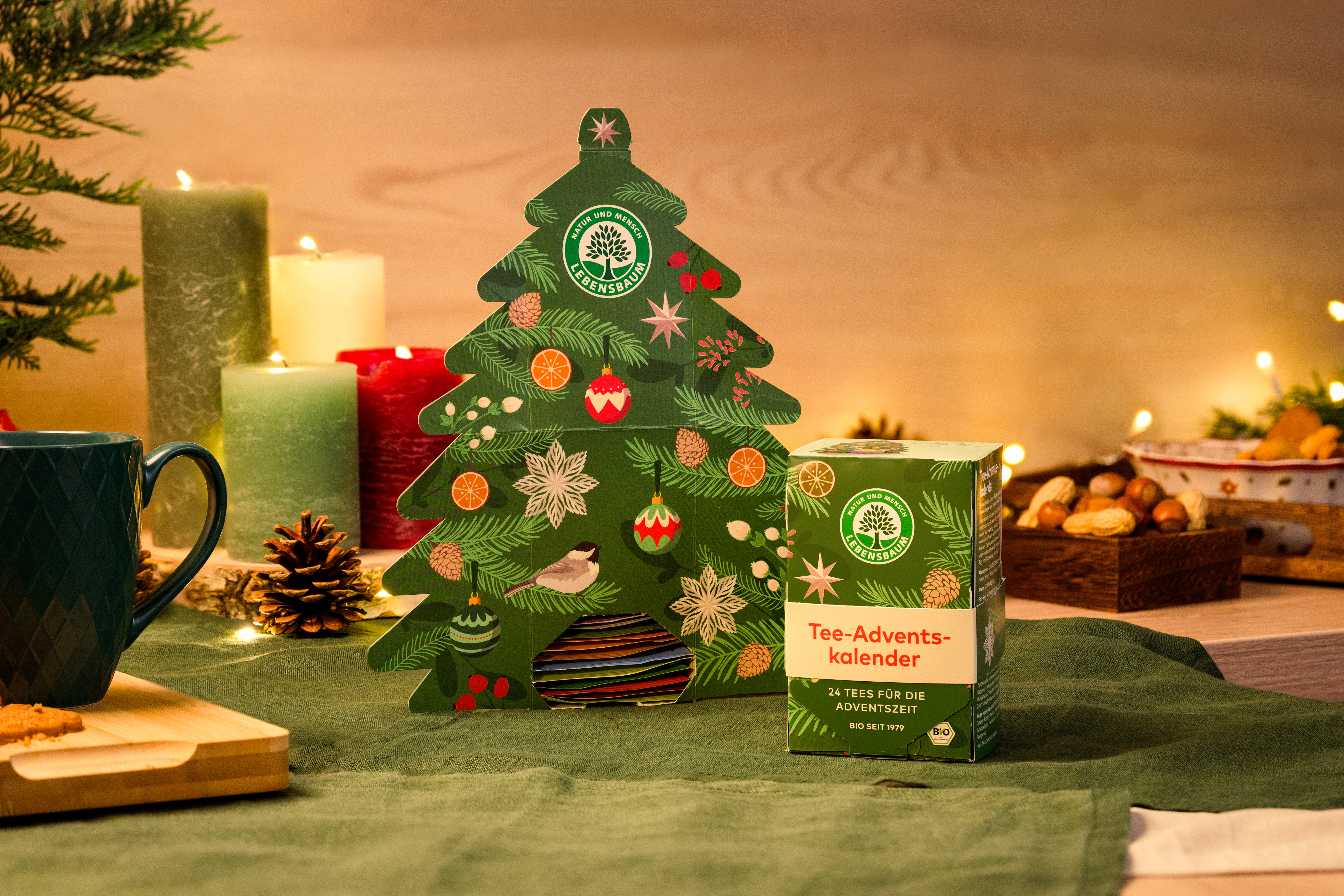 Tee-Adventskalender in grüner Weihnachtsbaumform neben brennenden Kerzen, Tannenzweigen und festlicher Dekoration auf grünem Untergrund.