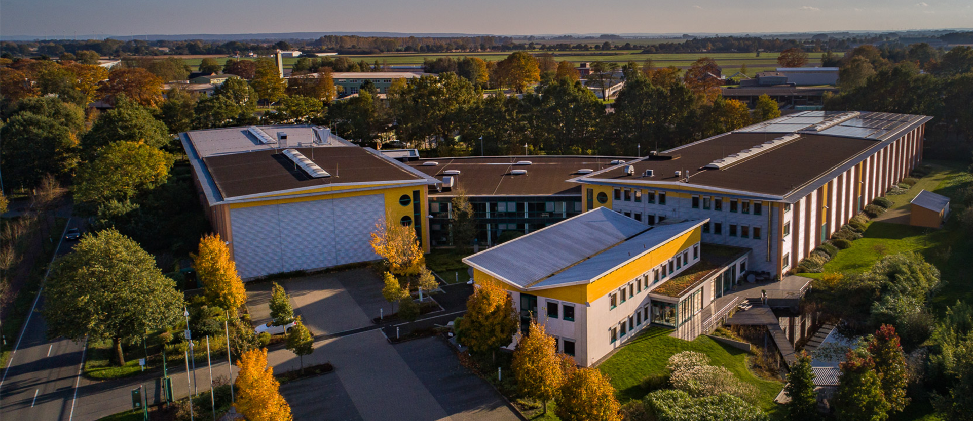 Moderne Produktionsfabrik mit gelb-weißen Gebäuden, Solarpanelen auf Dächern, umgeben von Herbstbäumen in ländlicher Umgebung.